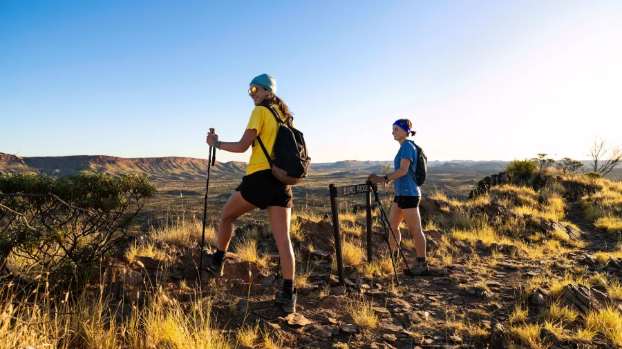 Two hikers with backpacks and walking poles stand on a ridge overlooking the rugged landscape of the Larapinta Trail.