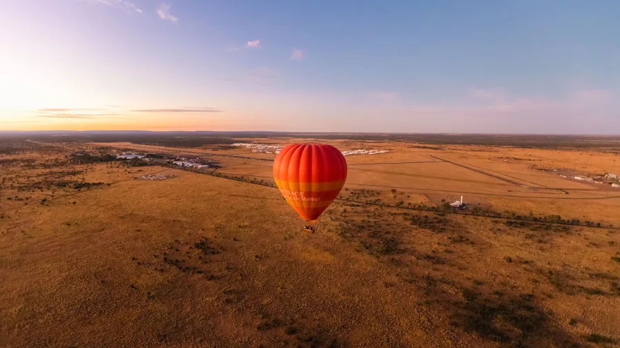 A hot air balloon floats above the MacDonnell Ranges at sunrise, overlooking a vast red and golden landscape.